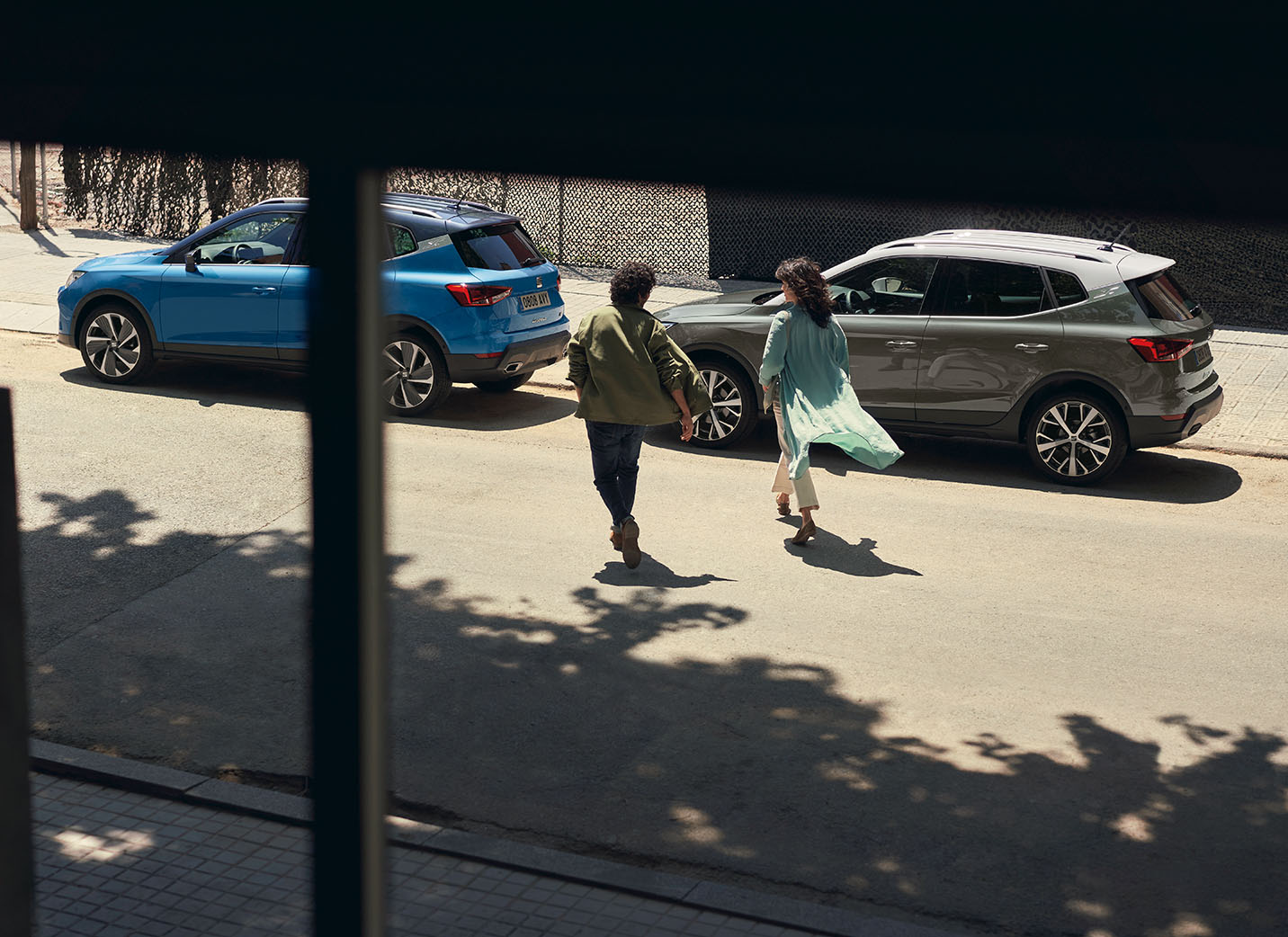 Sunny street scene where a man and a woman are walking towards two SEAT Arona 2024 cars, one blue and one grey parked along the curb. The setting suggests a casual, everyday urban environment