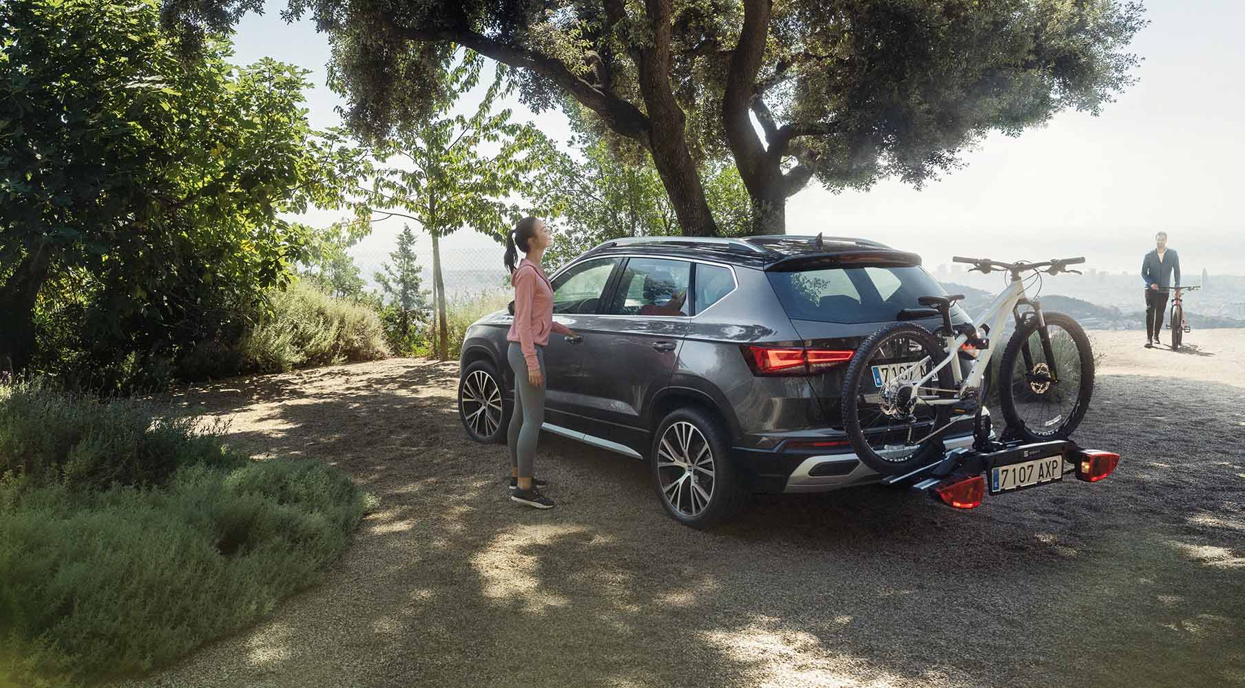 Sunlit scene of a dark grey SEAT Ateca 2024 equipped with a bike rack, parked on a gravel path by the coast. A man stands by the car, facing the ocean. Large trees and greenery surround the area, enhancing the serene coastal vibe.