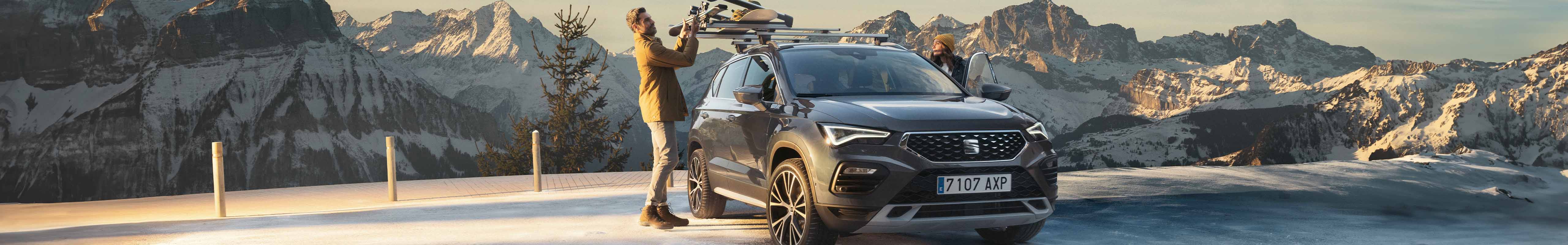A SEAT Ateca 2024 parked on a frost-covered road with a clear sky and rugged mountains in the background. Two people are visible, one standing beside the car and another with arms raised