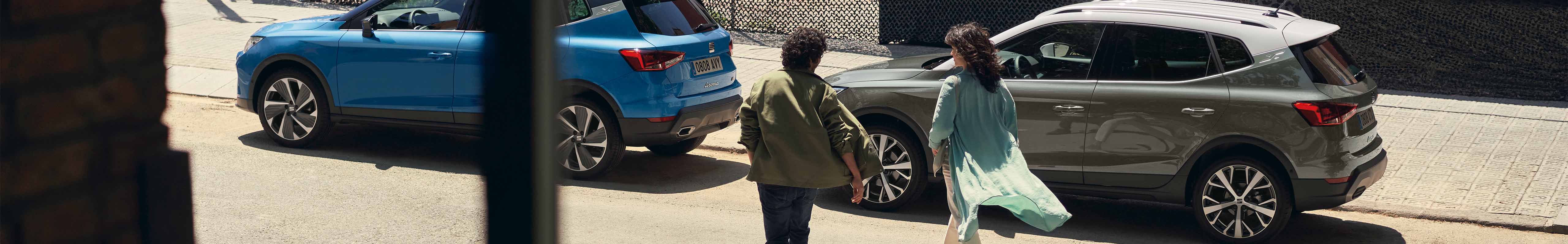 Sunny street scene where a man and a woman are walking towards two SEAT Arona 2024 cars, one blue and one grey parked along the curb. The setting suggests a casual, everyday urban environment.