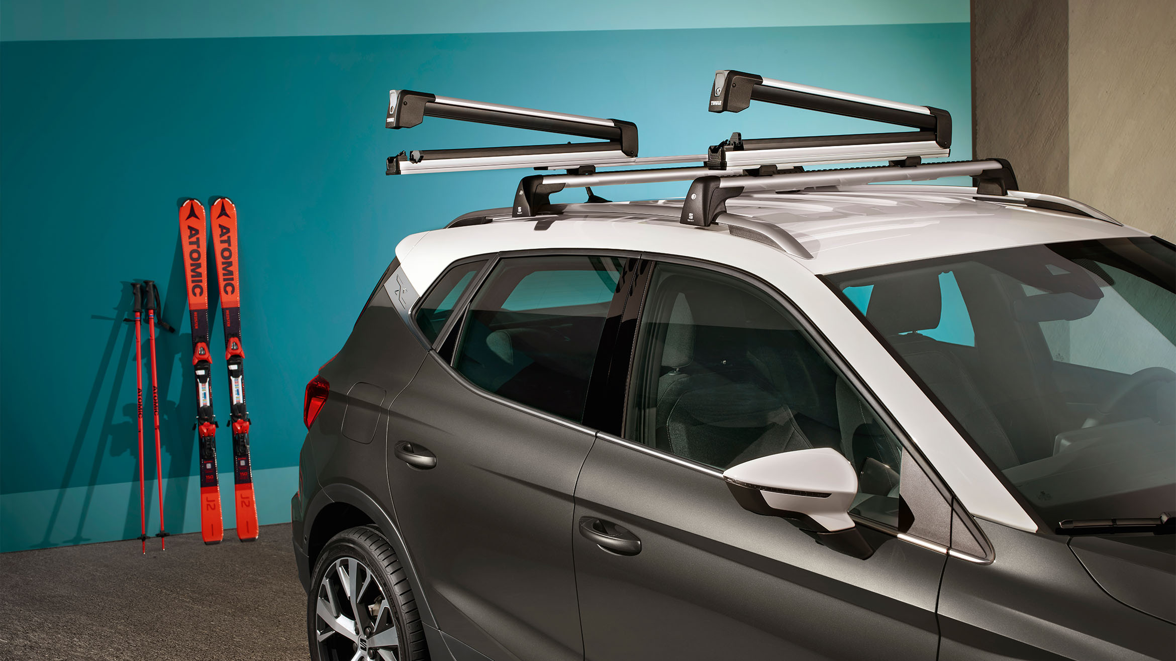 Rear view of a SEAT Arona in black with a white roof carrying skis in the ski rack xtender. The car is parked in a colourful setting outdoors