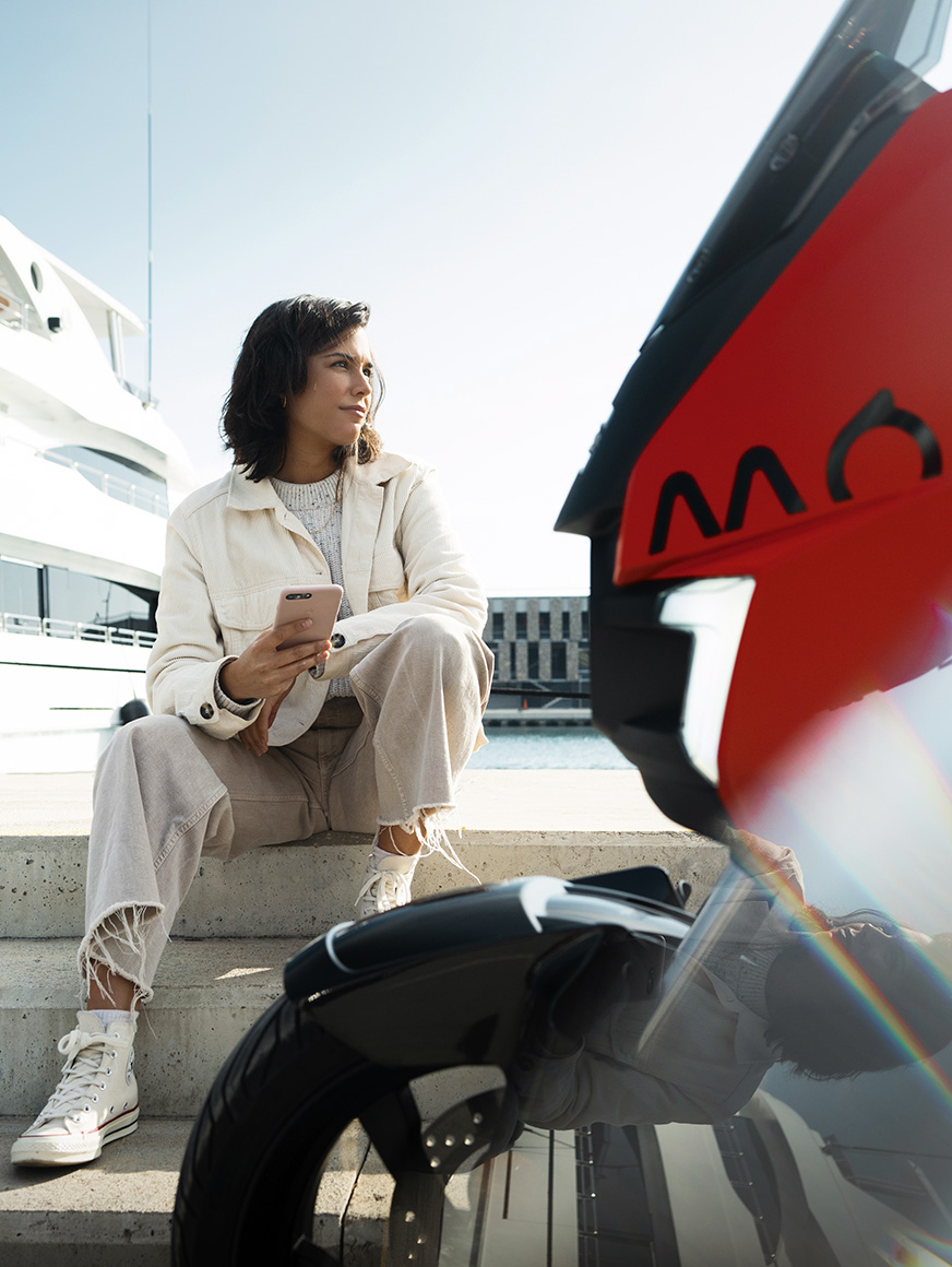 woman sitting by her parkedSEAT MÓ 125 electric scooter at a harbour