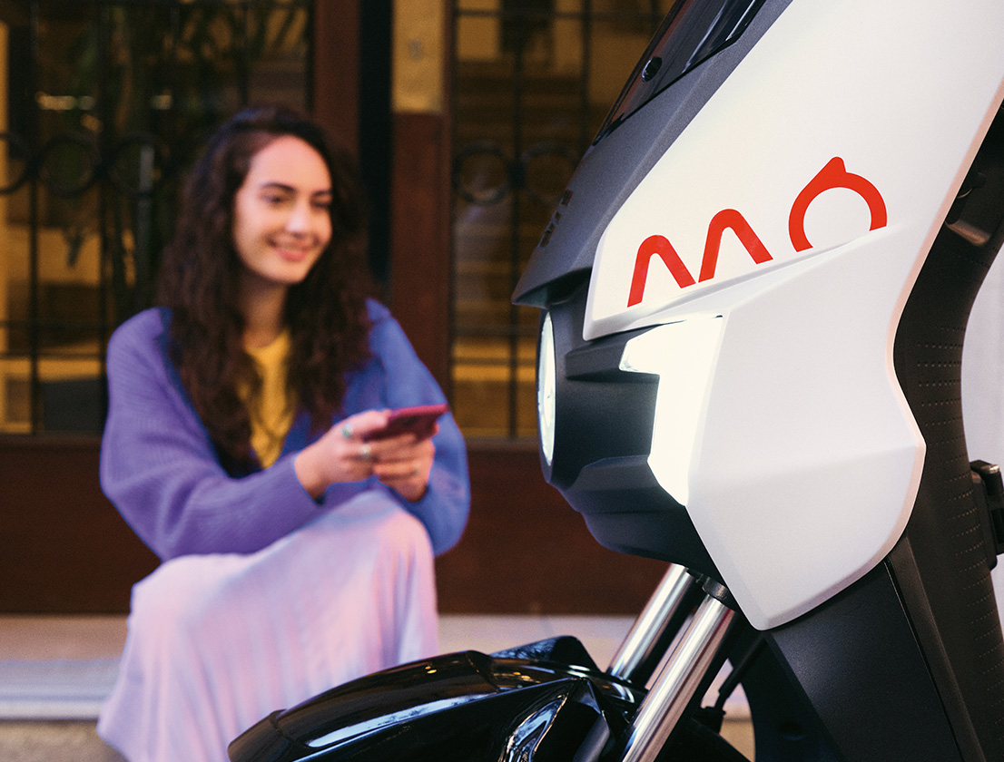  a girl sitting in frotn of her SEAT MO 125 scooter with the smart front light on