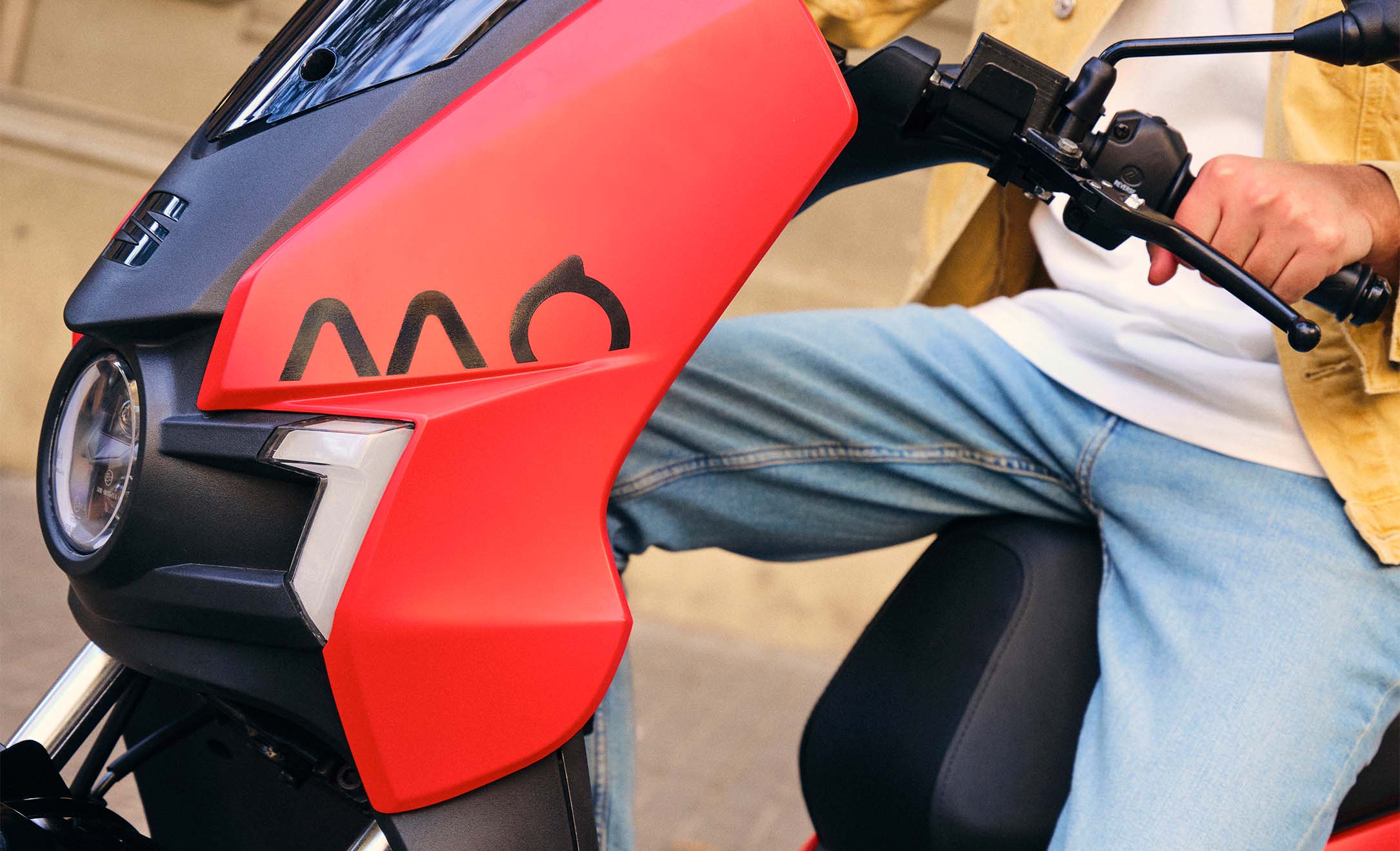Close up view of a seat mo 125 highlighting the red colour of this model, the sleek design details of the electric scooter and the seat mo logo. It is being driven by an individual dressed in jeans.