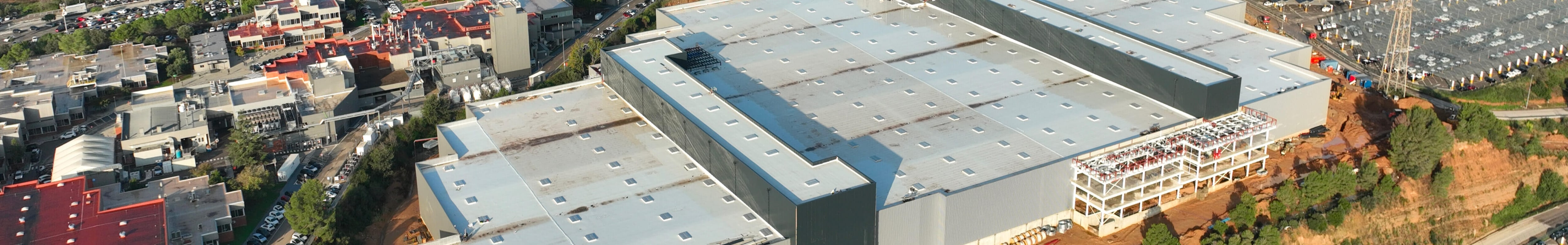 An aerial shot showcases a sprawling factory complex with multiple large buildings and extensive rooftops and roadways under a partly cloudy sky.