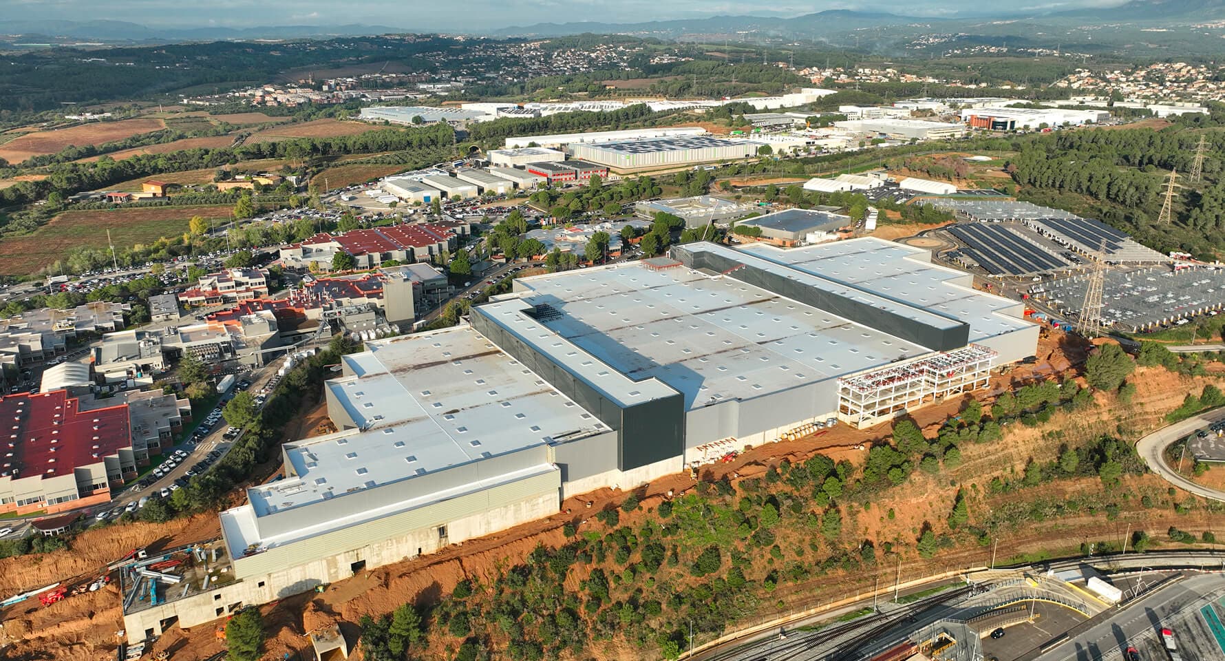 An aerial shot showcases a sprawling factory complex with multiple large buildings and extensive rooftops and roadways under a partly cloudy sky.