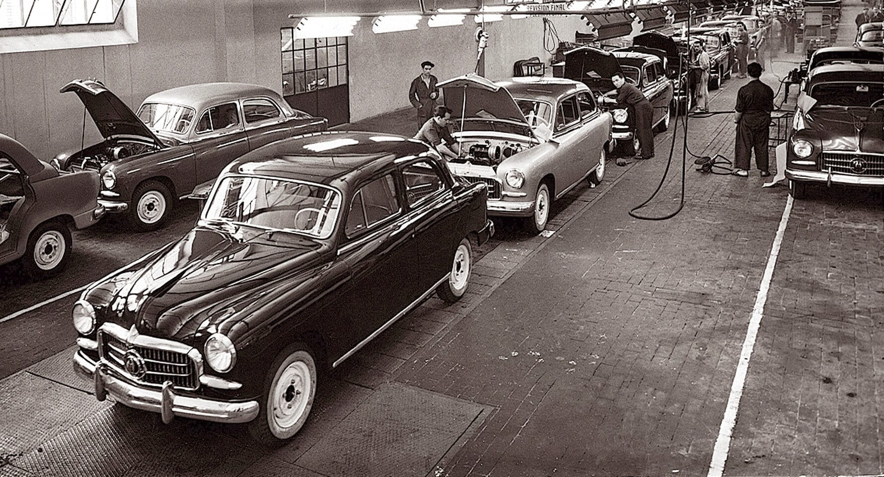Vintage car assembly line with workers and classic cars in a factory.