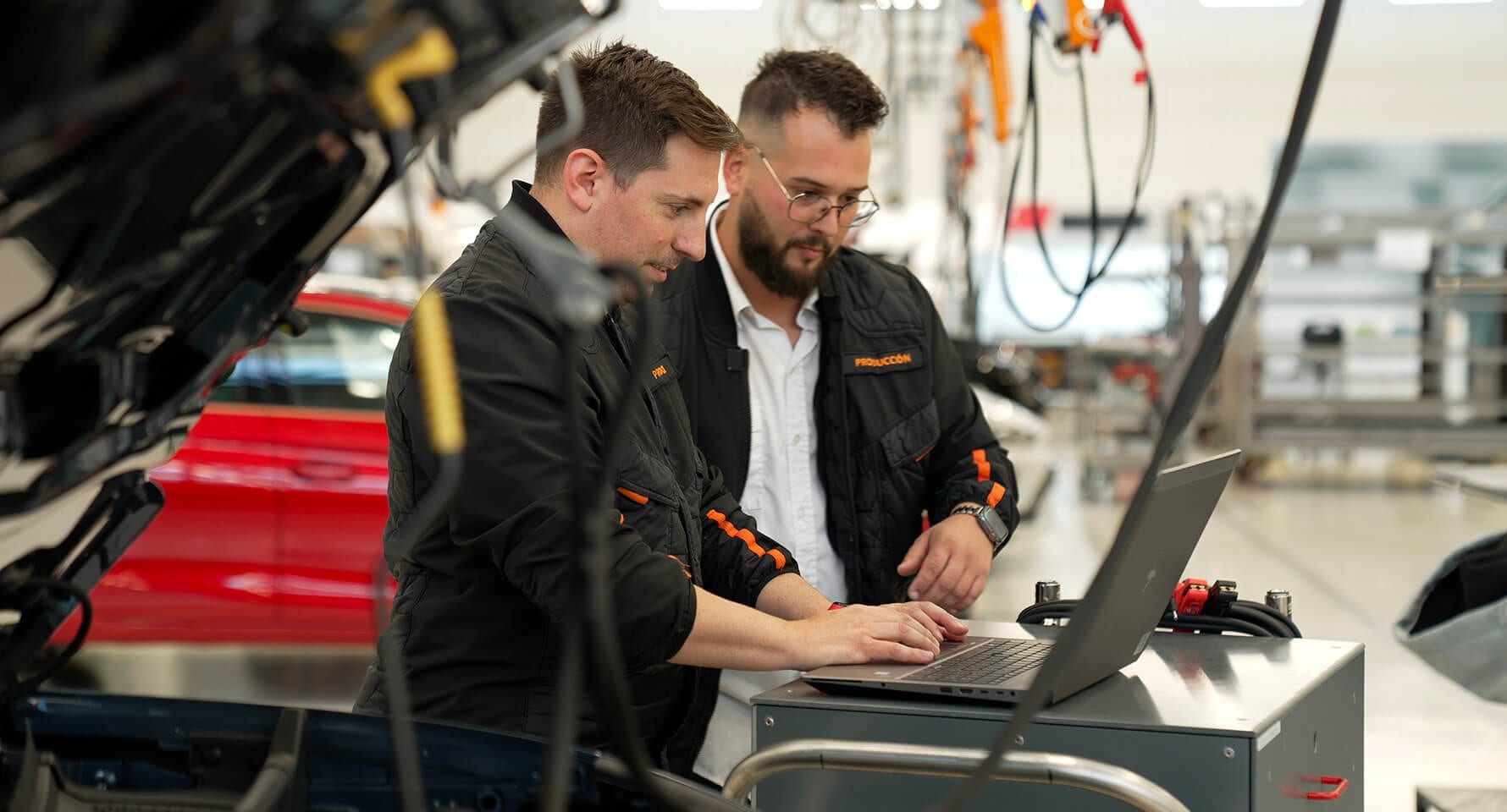 Two men wearing black jackets with orange accents are looking at a laptop connected to a car with its hood open in a workshop or testing facility.