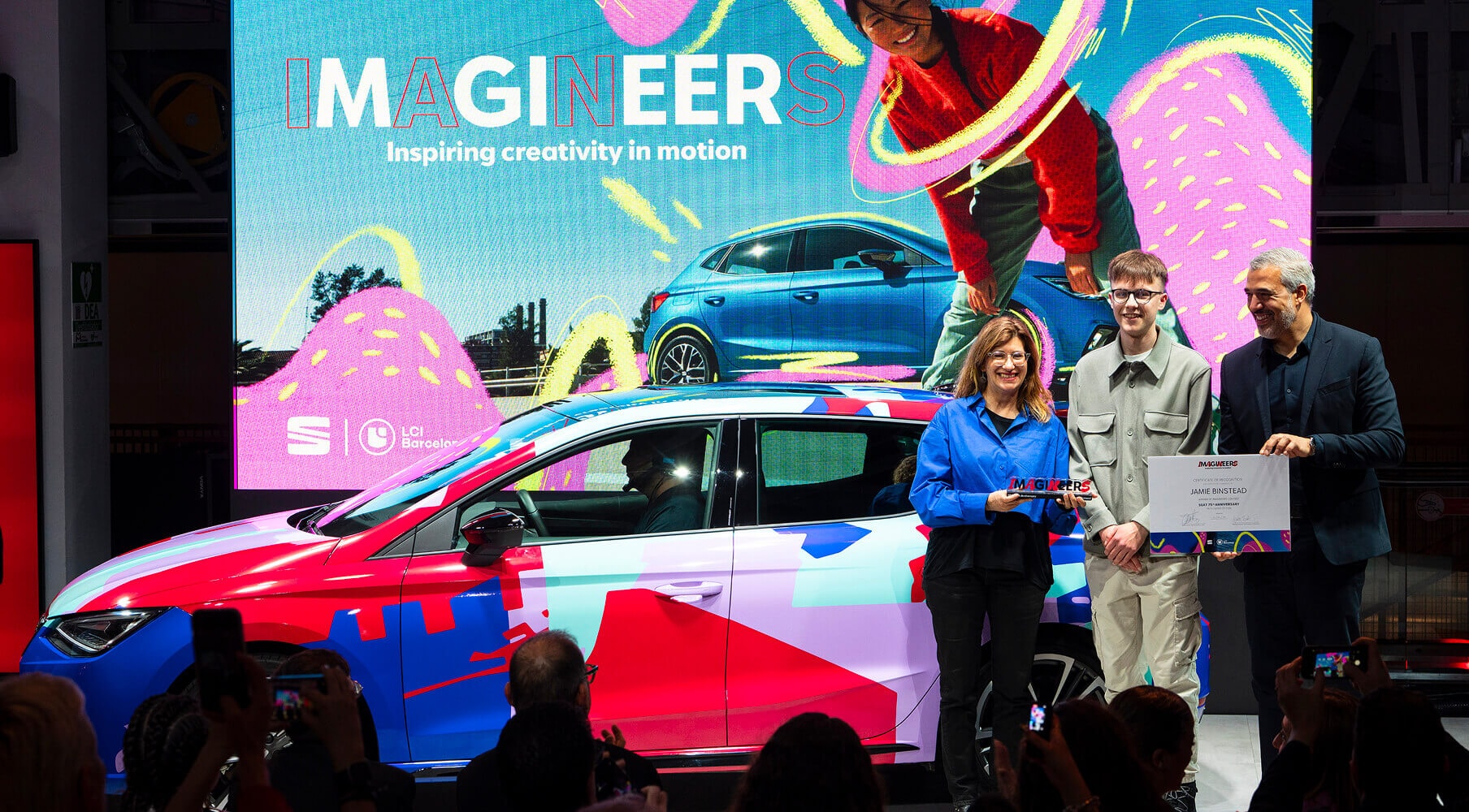 The winner of the SEAT Imagineers design competition receiving an award, standing next to Francesca Sangalli and Jorge Diez. In the background is the winning car design, decorated with a colourful, abstract pattern.