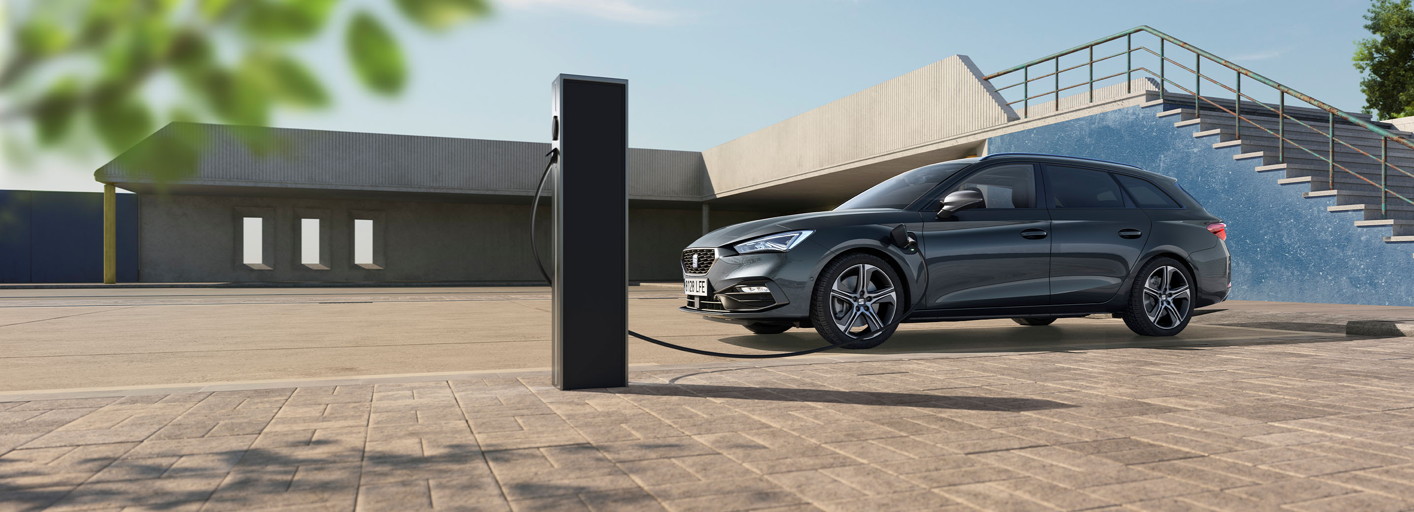 A SEAT Leon Sportstourer parked on a sunlit paved plaza, connected via a charging cable to a pedestal station. The background features modern concrete architecture and an outdoor staircase with a vibrant blue wall under a clear sky.