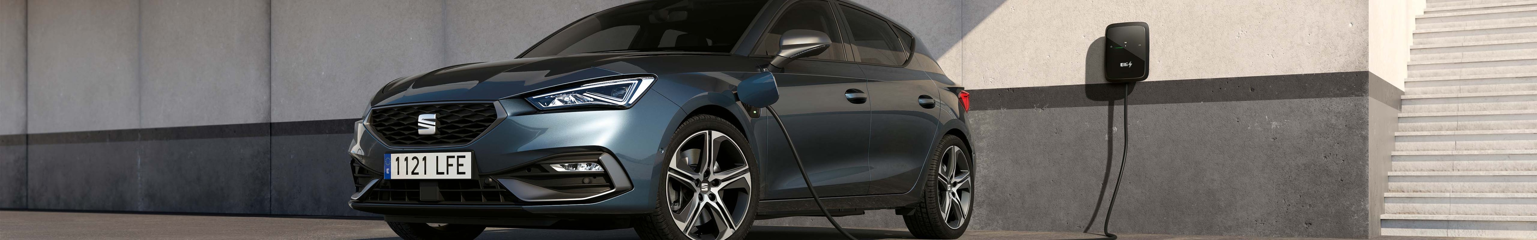 A Magnetic Tech Grey SEAT Leon hybrid parked beside a concrete wall, with a visible cable connecting it to a wallbox charger.