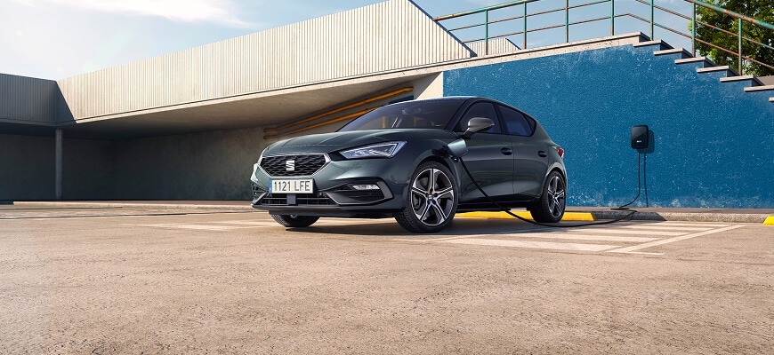 Hero front angle view of a Magnetic Tech Grey SEAT Leon e-HYBRID FR connected to a Wallbox charger, parked in front of a modern building.