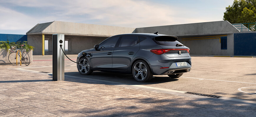 Rear and side view of a Magnetic Tech Grey SEAT Leon Sportstourer e-HYBRID parked near a public charging point, with a modern building and trees in the background.