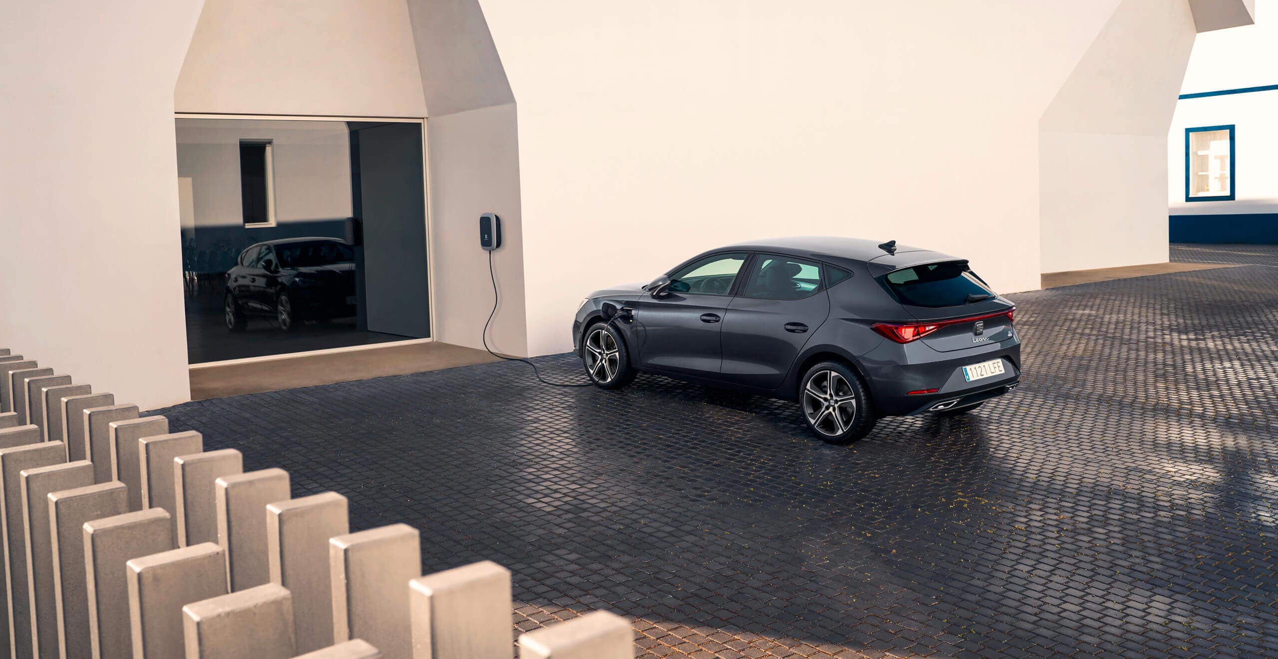 A SEAT Leon e-HYBRID in Magnetic Tech Grey is charging, parked beside a modern building with reflective windows. 