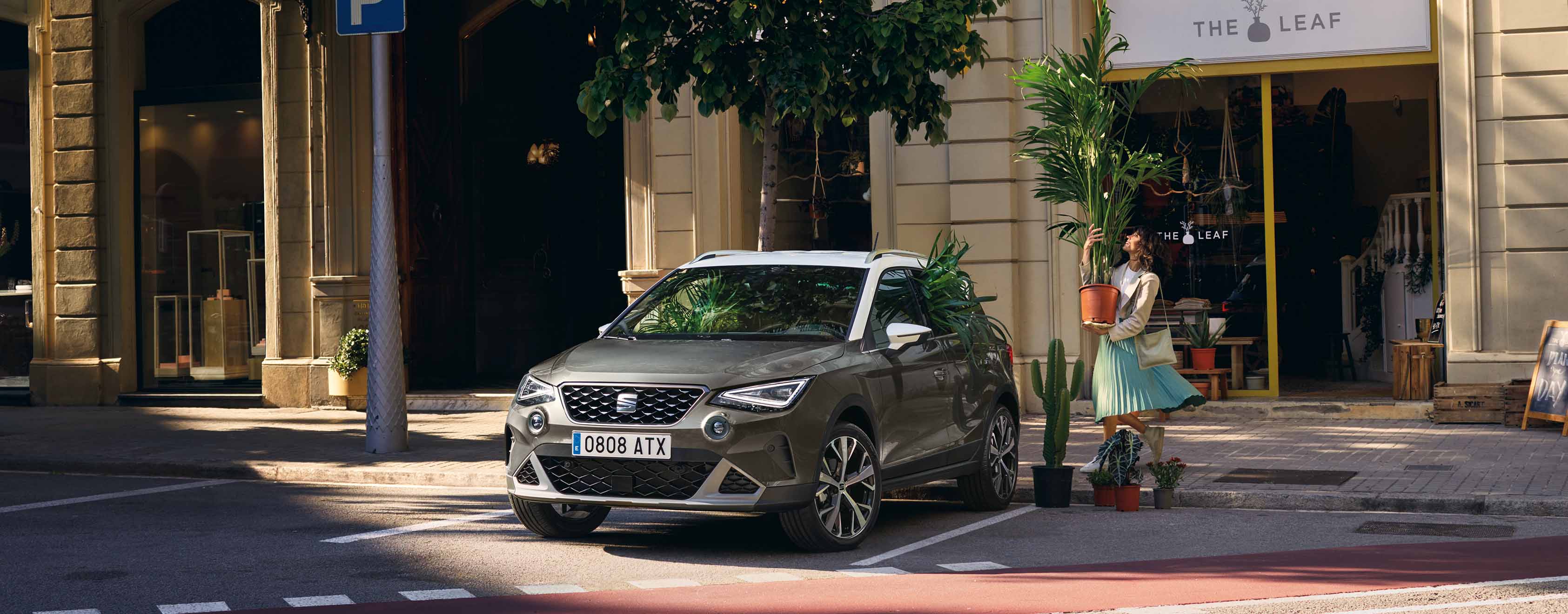 A silver seat arona 2024 suv parked along a shaded city street, with sunlight filtering through trees. The background features a historic building with arches and a small boutique shop. The scene captures a peaceful urban morning