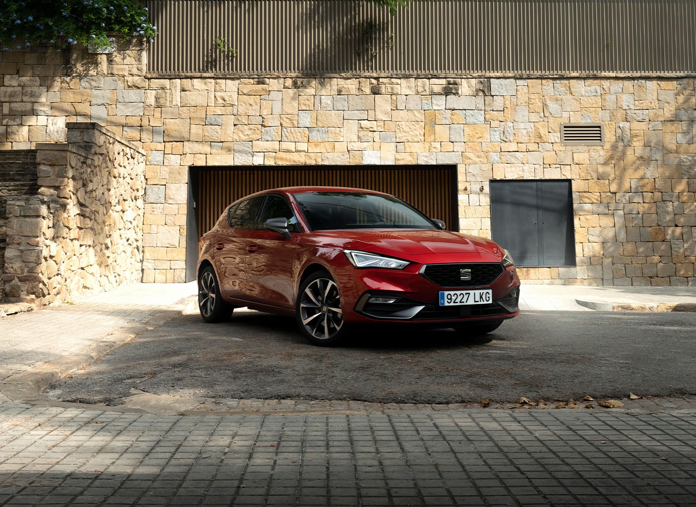A red SEAT Leon hatchback is parked in a driveway in front of a stone wall garage opening. The setting is suburban, with lush green trees and a modern fence above the stone wall.