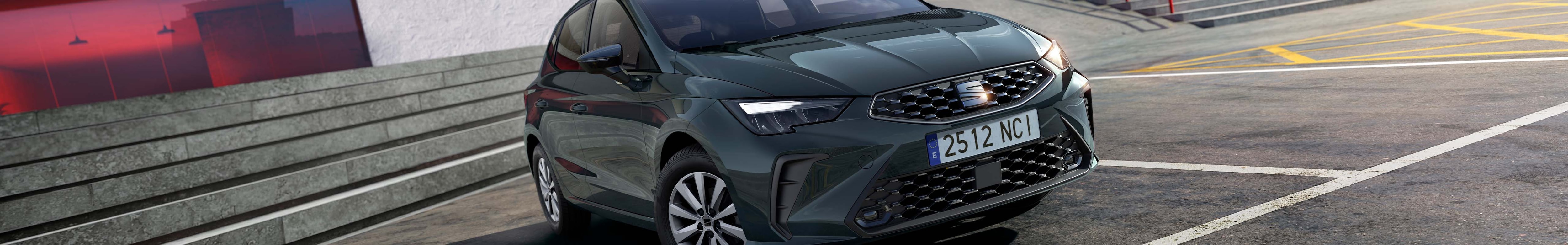 A SEAT Ibiza is parked in an asphalt lot with yellow and white markings. In the background is a striking, modern building with sharp angles in white and red, including a prominent staircase.