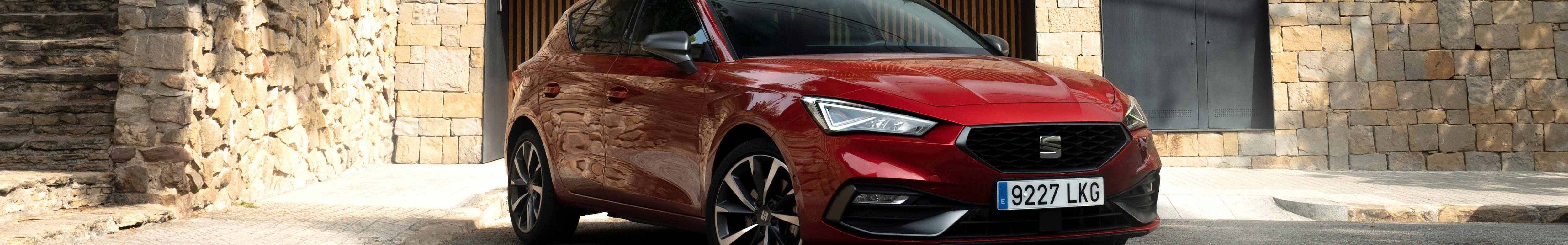 A red SEAT Leon hatchback is parked in a driveway in front of a stone wall garage opening. The setting is suburban, with lush green trees and a modern fence above the stone wall.