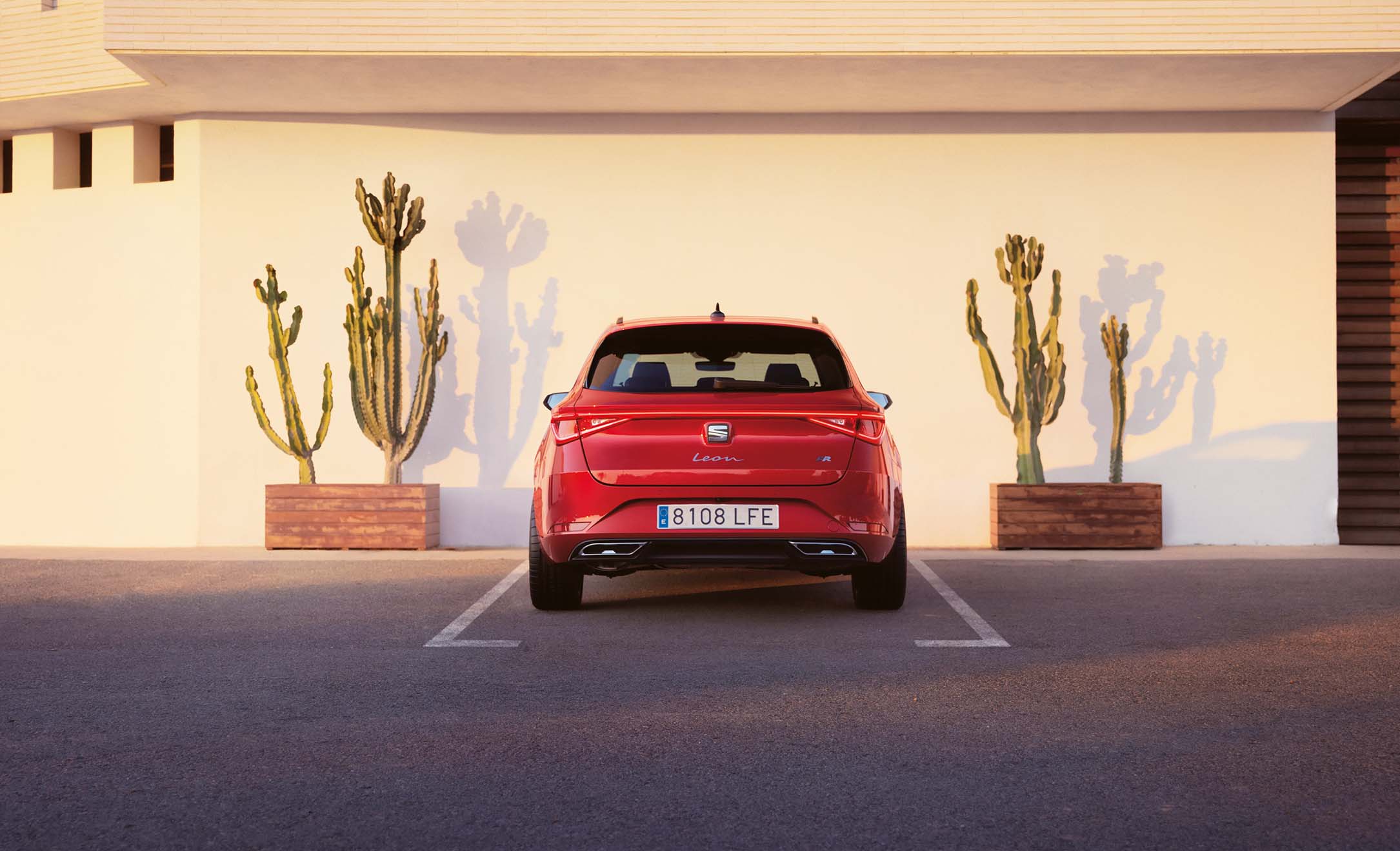 Rear view of a red SEAT Leon Sportstourer 2024 in a car park, with desert-like landscaping including tall cacti in the background 