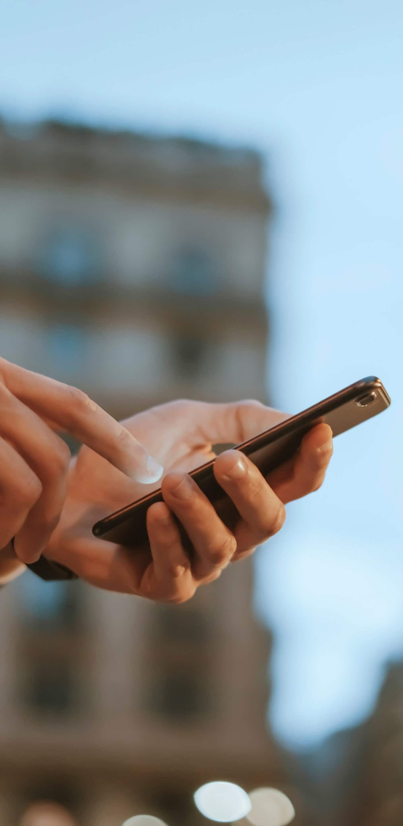 A close-up shot of a person's hands holding and interacting with a smartphone on a street. Blurred city lights and buildings are visible in the background.