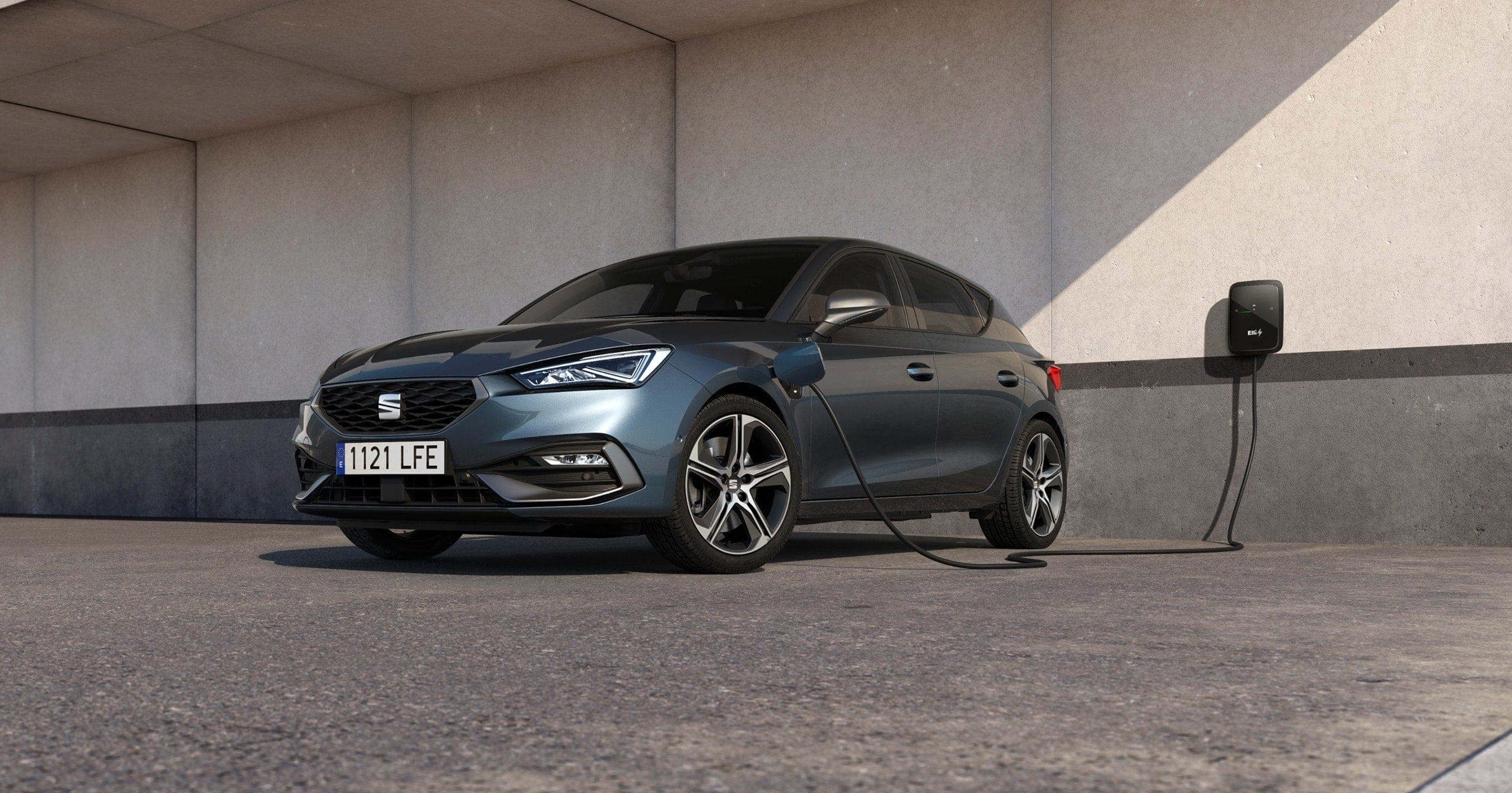 A dark gray SEAT Leon compact car is plugged into a wall charging station in a modern, concrete outdoor space. A wide staircase is visible on the right.